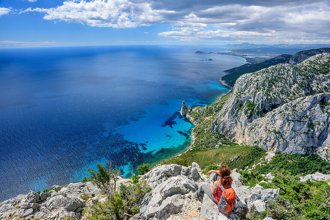 Woman hiking Selvaggio Blu sitting at Punta Giradili and looking at coast of Golfo di Orosei with rock spire Pedra Longa, Selvaggio Blu, National Park of the Bay of Orosei and Gennargentu, Sardinia, Italy