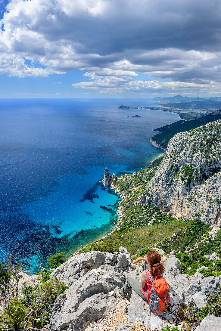 Frau beim Wandern sitzt an Punta Giradili und blickt auf Golfo di Orosei mit Felsnadel Pedra Longa, Selvaggio Blu, Nationalpark Golfo di Orosei e del Gennargentu, Sardinien, Italien