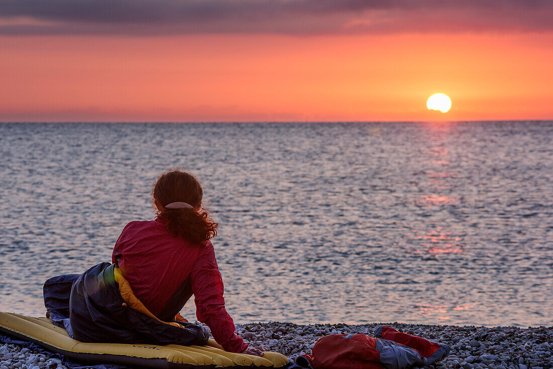 Frau liegt in Schlafsack am Strand und blickt auf Sonnenaufgang, Selvaggio Blu, Nationalpark Golfo di Orosei e del Gennargentu, Sardinien, Italien