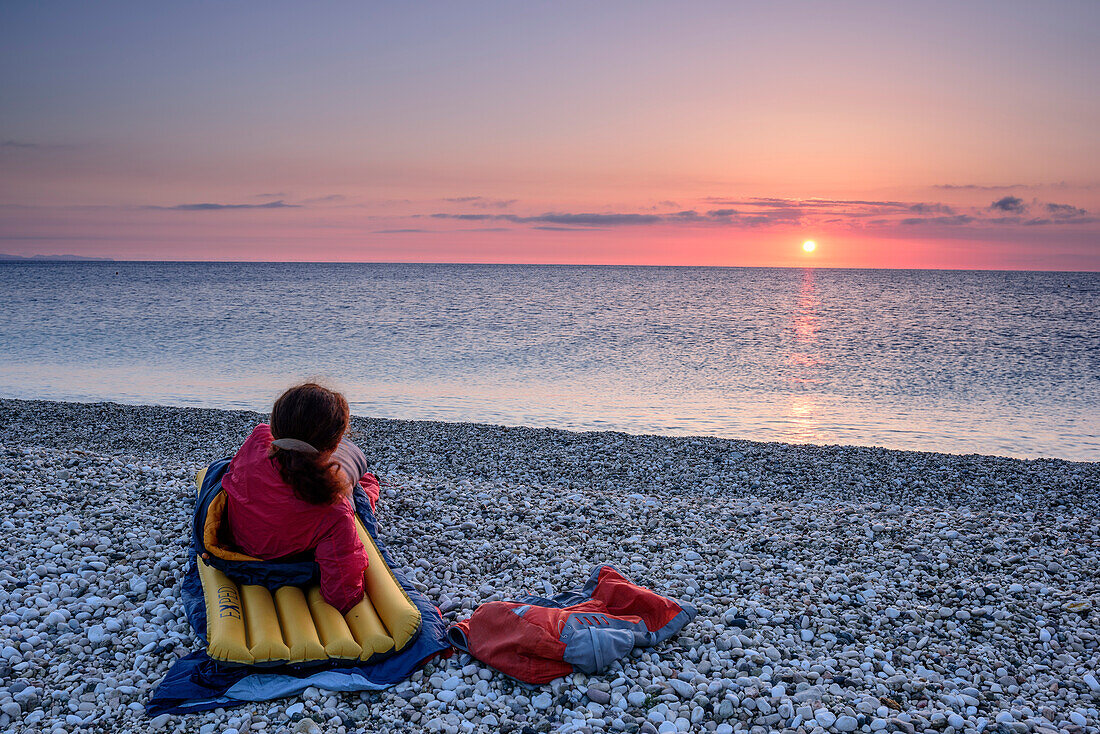 Frau liegt in Schlafsack am Strand und blickt auf Sonnenaufgang, Selvaggio Blu, Nationalpark Golfo di Orosei e del Gennargentu, Sardinien, Italien