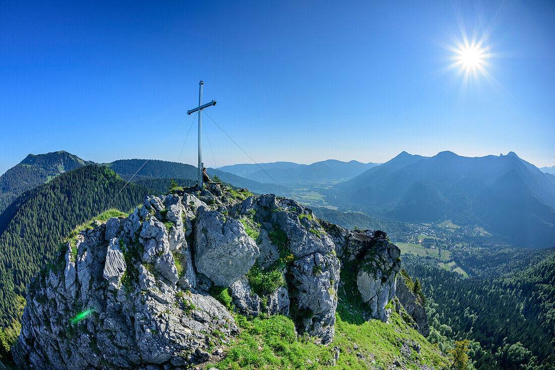 Person sitzt am Gipfelkreuz des Leonhardstein, Blick auf Tegernseer Berge, Leonhardstein, Bayerische Alpen, Oberbayern, Bayern, Deutschland