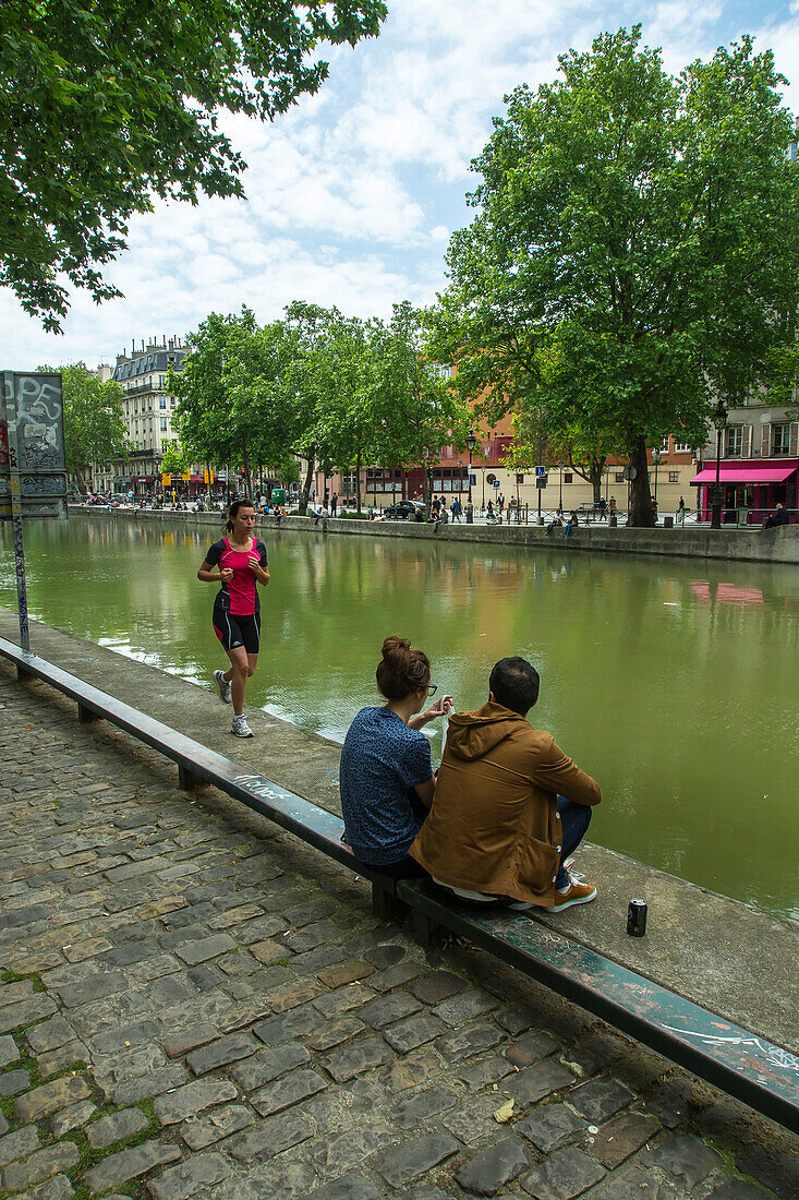 France, Paris, Jogger woman and a couple sitting on the edge of Canal St Martin