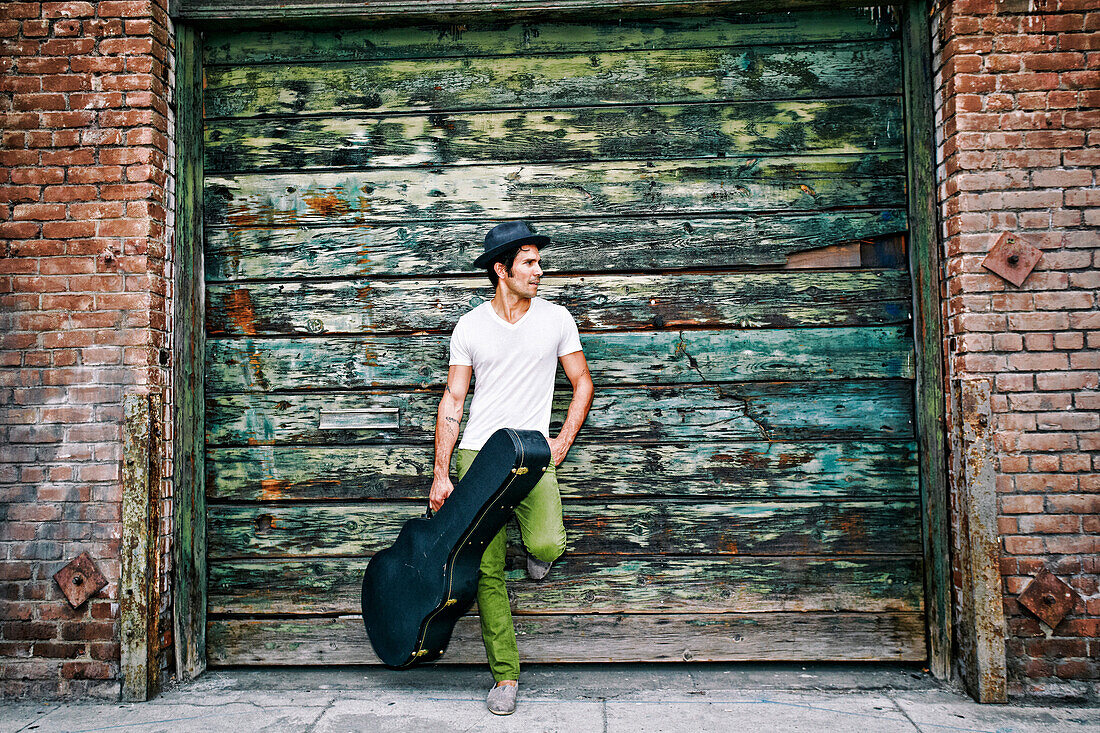 Mixed race musician holding guitar case near industrial building, Los Angeles, California, USA