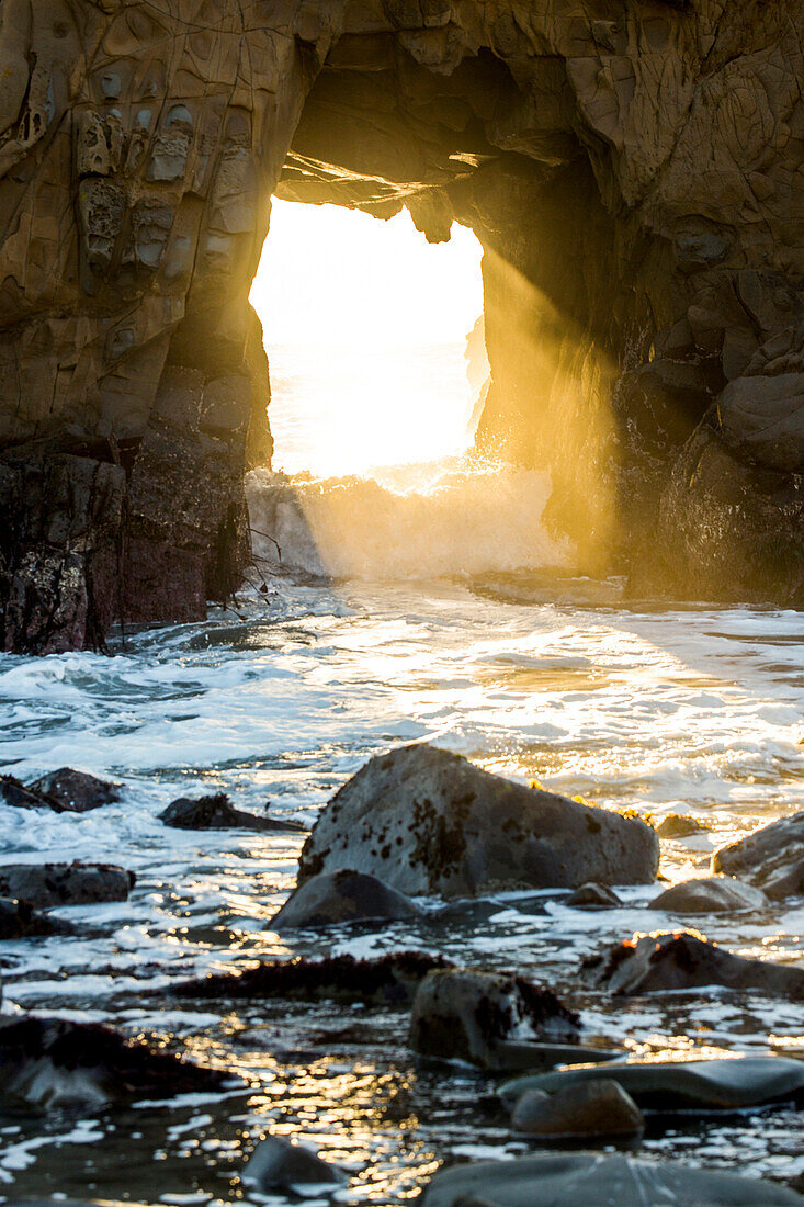 Sunbeam shining through rock formation to ocean waves, Big Sur, California, United States