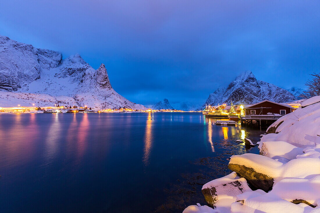 Blue hour in the small bay of Reine, where the lights of the houses reflect in the calm water surrounding the Lofoten Islands, Arctic, Norway, Scandinavia, Europe