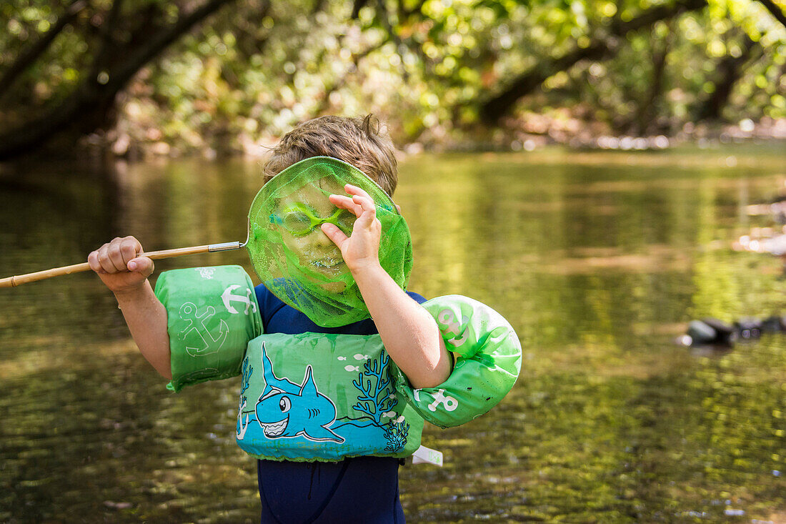 Toddler boy with floatie and goggles spies camera through net in creek at Bidwell Park, Chico, California.
