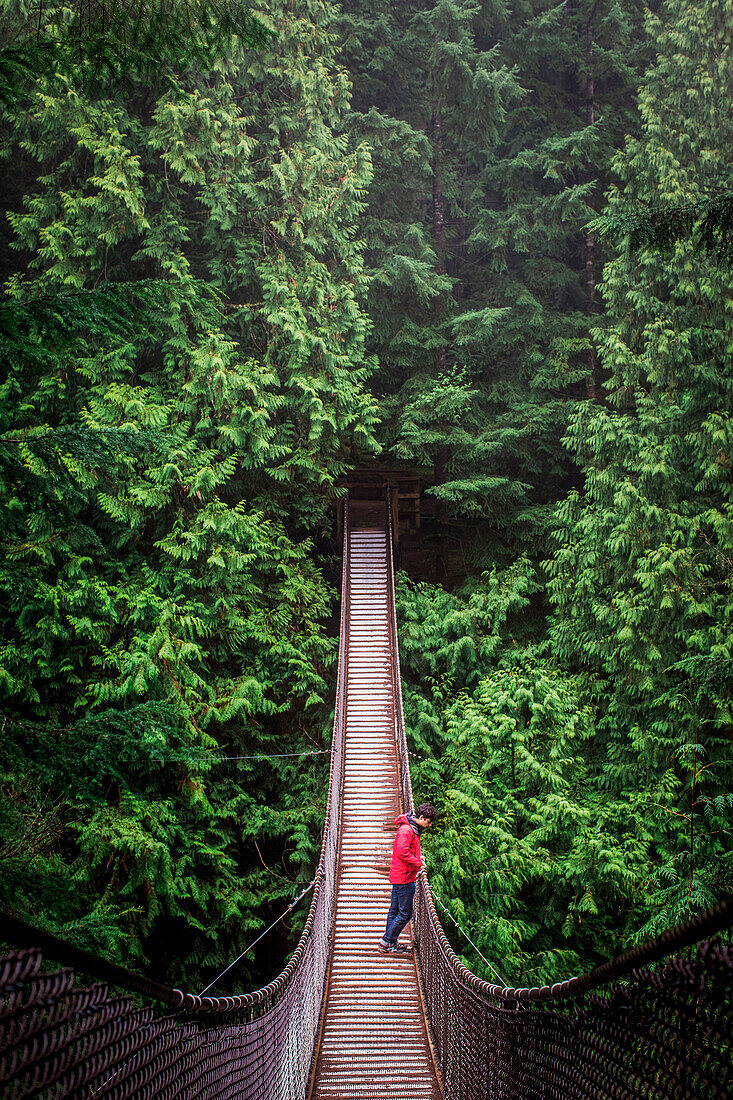 A man in a red coat and jeans stands in middle of a suspension bridge surrounded by large green trees.