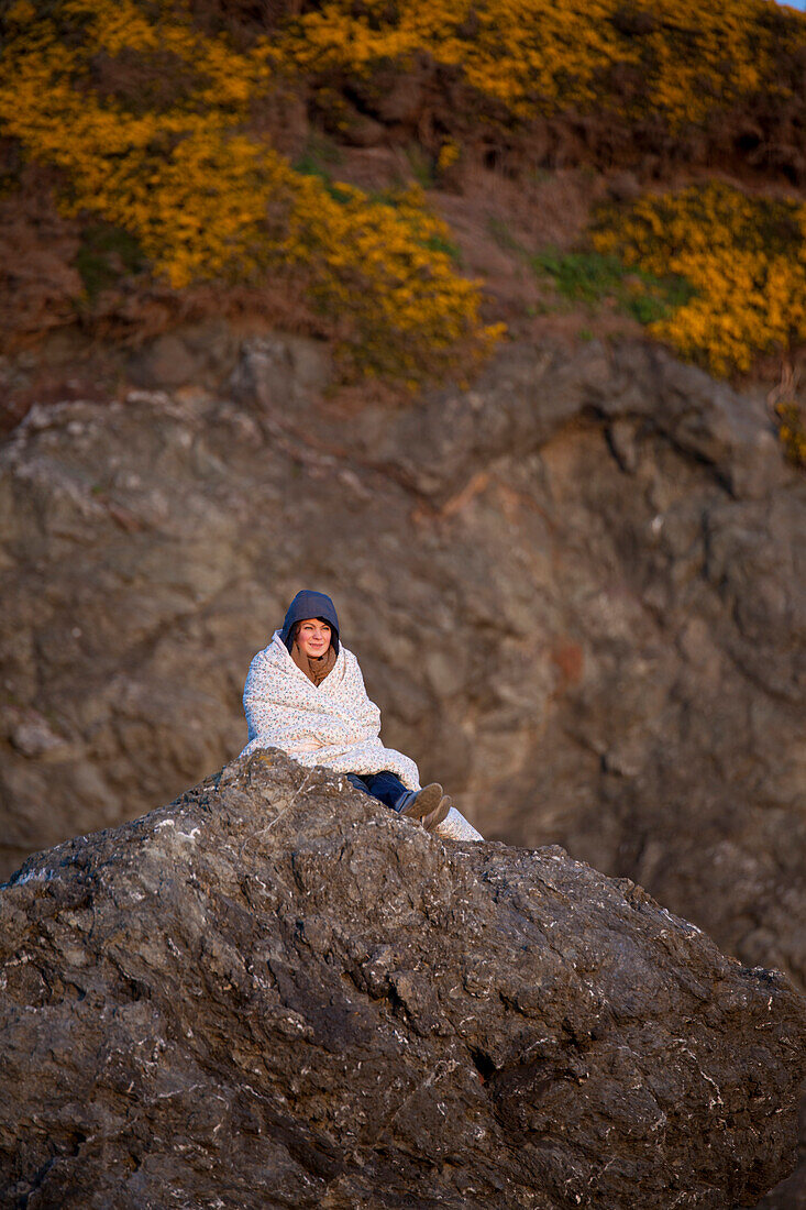 A young woman wrapped in a blanket sists on a rock bluff at Bandon Bay, Oregon.