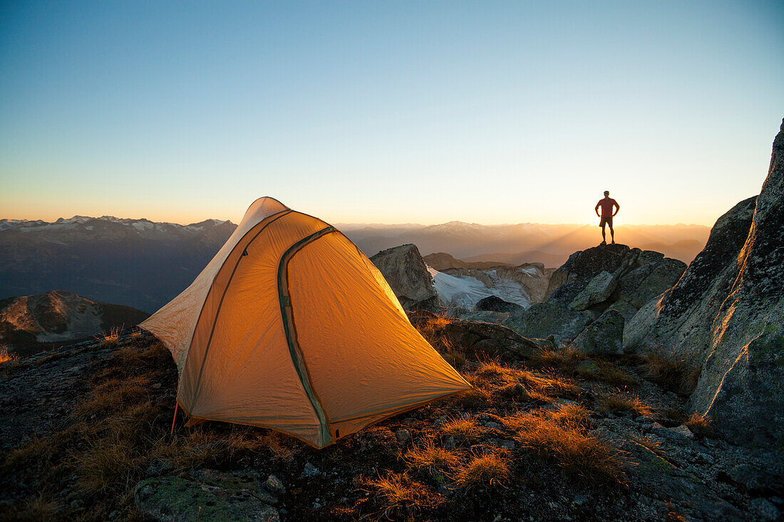 A silhouetted hiker stands on a rock bluff while camping on the summit of Saxifrage Peak, Pemberton, BC, Canada.