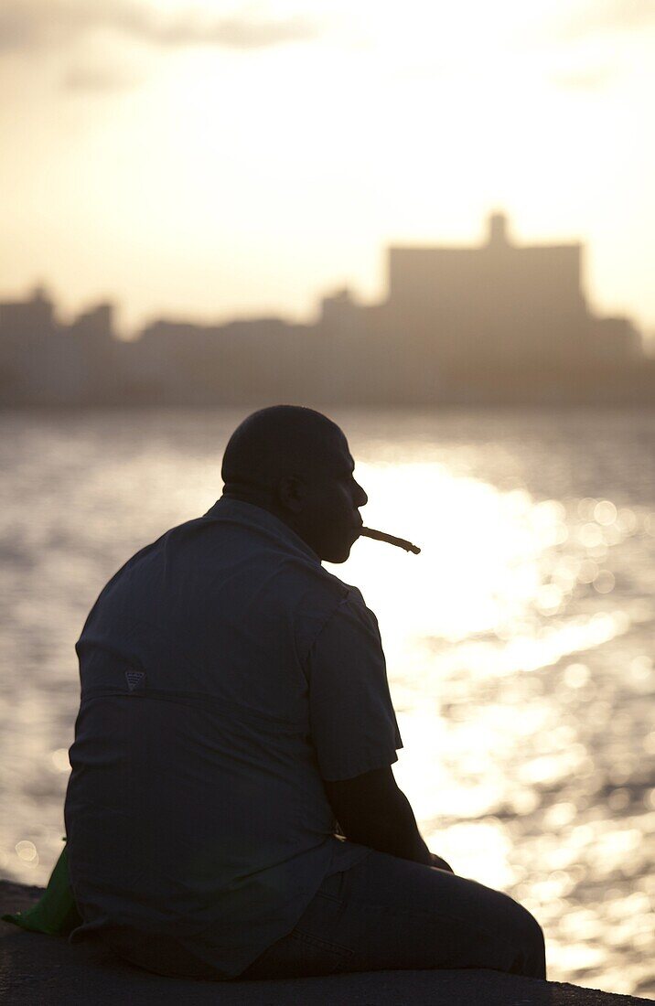 Man in semi silhouette sat on the sea wall at sunset, smoking a cigar, The Malecon, Havana Centro, Havana, Cuba, West Indies, Central America