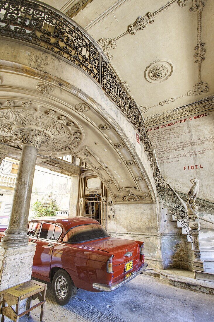 Classic red American car parked beneath ornate marble staircase inside dilapidated apartment building, Havana, Cuba, West Indies, Central America