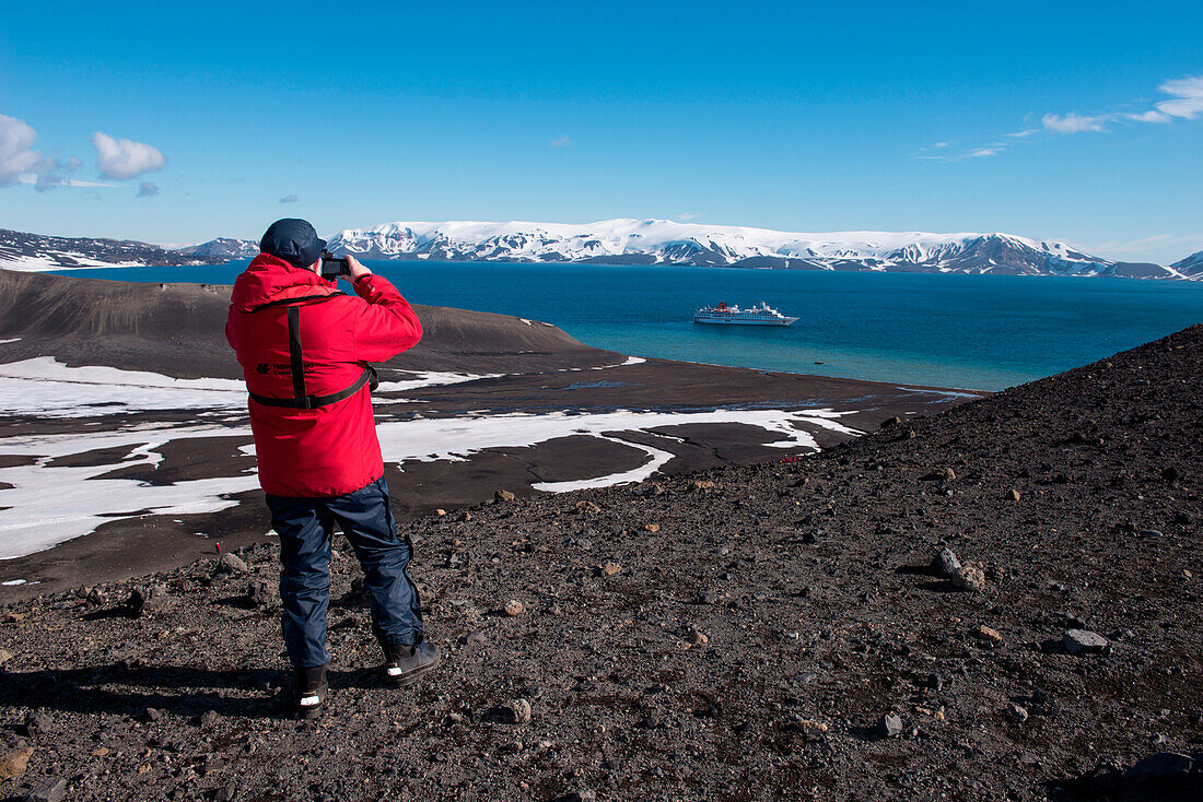 Man in red expedition parka stands at crater edge and takes a photograph of expedition cruise ship MS Hanseatic (Hapag-Lloyd Cruises), Telephone Bay, Deception Island, South Shetland Islands, Antarctica