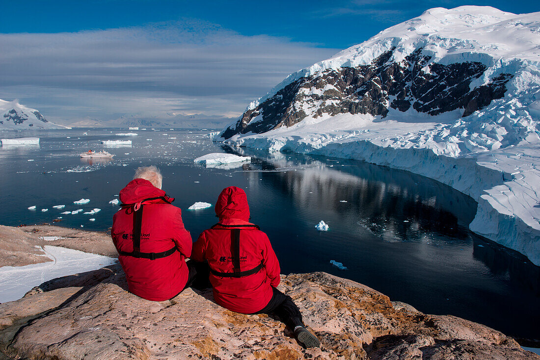 Passengers from expedition cruise ship MS Hanseatic (Hapag-Lloyd Cruises) sit on rock and admire view from hilltop, Neko Harbour, Graham Land, Antarctica