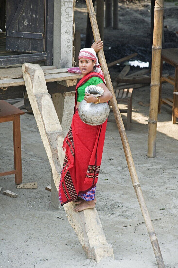 Assamese tribal village woman with water pot, climbing simple wooden steps up to her house, Majuli Island, Assam, India
