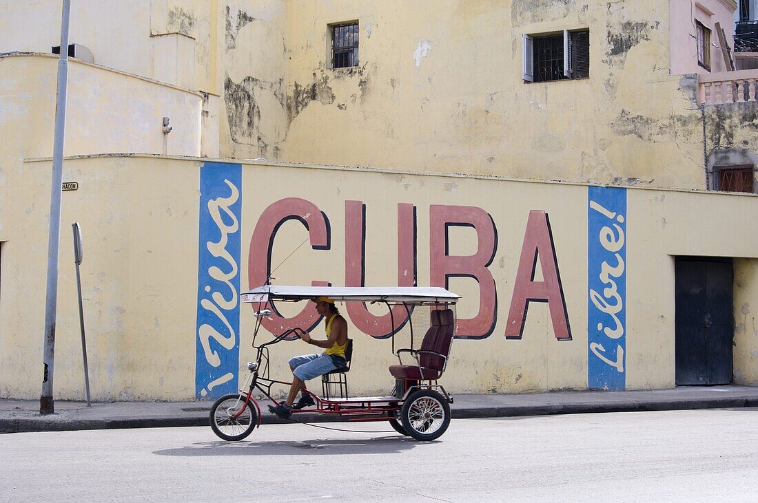 A rickshaw passing a wall sign celebrating Cuban freedom, Havana, Cuba, West Indies, Central America
