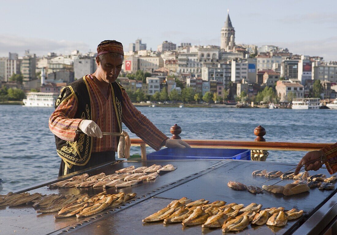 Man cooking fish on a  boat on the Golden Horn with the Galata Tower and Beyoglu district in the background, Istanbul, Turkey, Europe