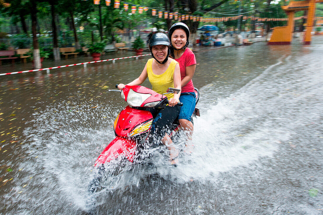 Zwei fröhliche Frauen fahren mit Moped durch Wasser von überfluteter Straße, Ho-Chi-Minh-Stadt (Saigon), Ho Chi Minh, Vietnam, Asien