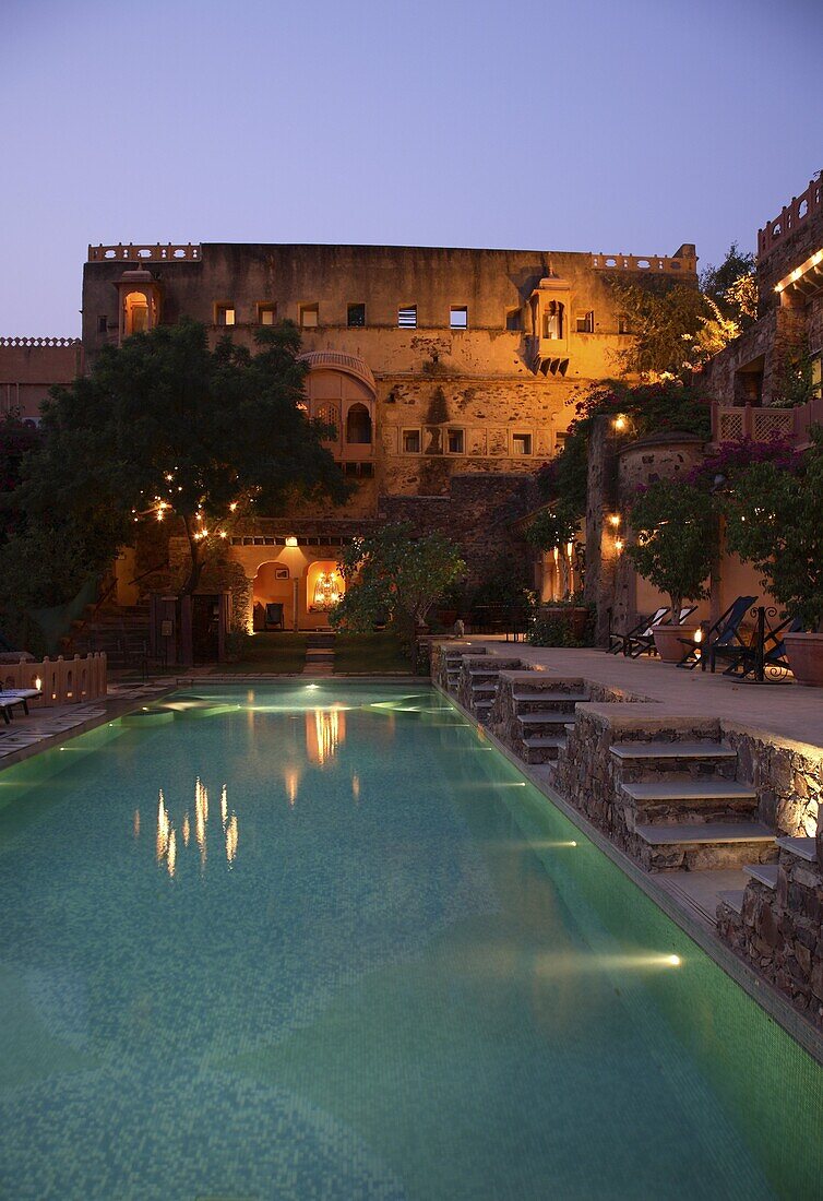 Rough-hewn walls with balconies, arches, and windows rise up on two sides of the beautifully-lit swimming pool, Neemrana Fort Palace, Rajasthan, India, Asia