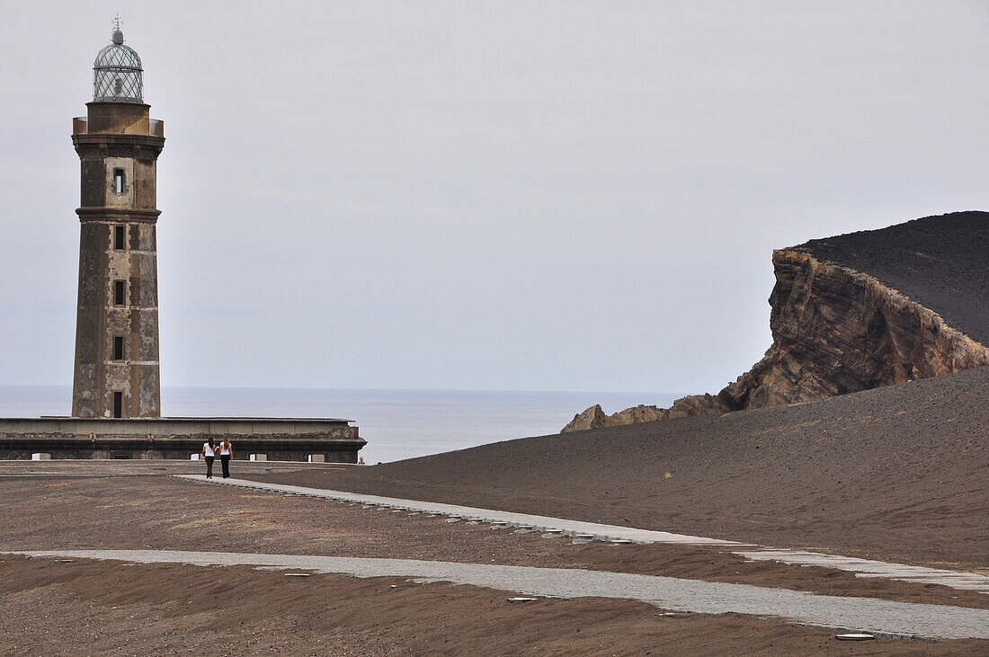 Two young woman hiking at Lighthouse at beach with volcano Vulcao dos Capelinhos, Farol dos Capelinhos, Ponta dos Capelinhos, Island of Faial, Azores, Portugal, Europe, Atlantic Ocean