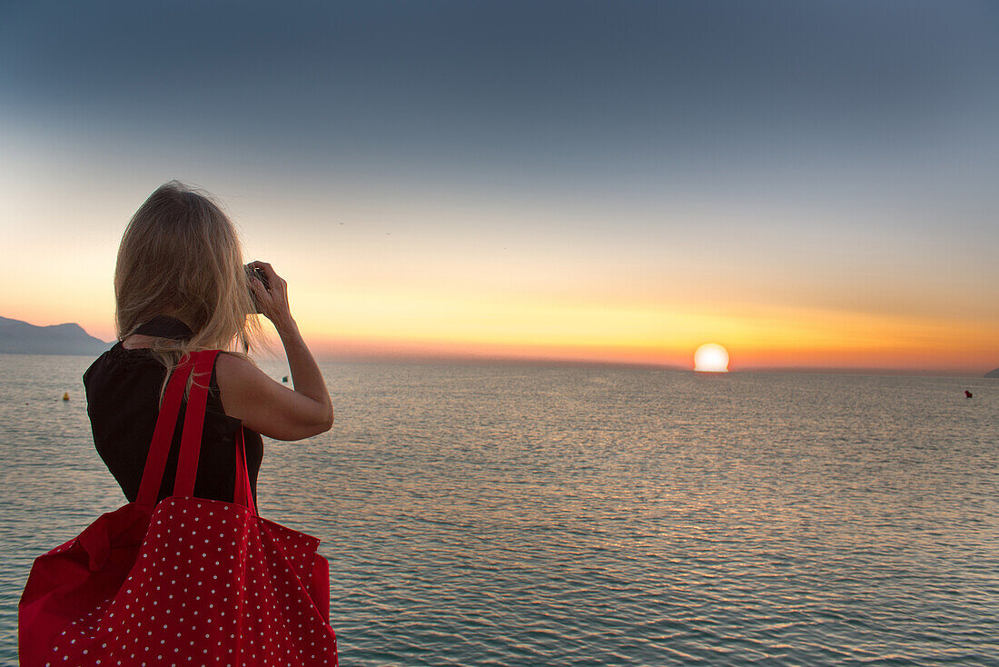 Eine Frau mit roter Tasche fotografiert den Sonnenaufgang am Mittelmeer. Strand Playa de Muro, Alcudia, Mallorca, Balearen, Spanien
