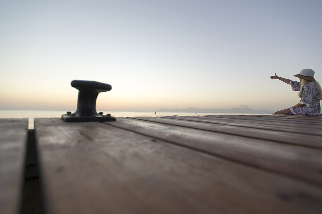 Junge Frau sitzt am Ende eines langen Steg in der Morgenstimmung und zeigt mit der Hand auf das Mittelmeer. Strand Playa de Muro, Alcudia, Mallorca, Balearen, Spanien