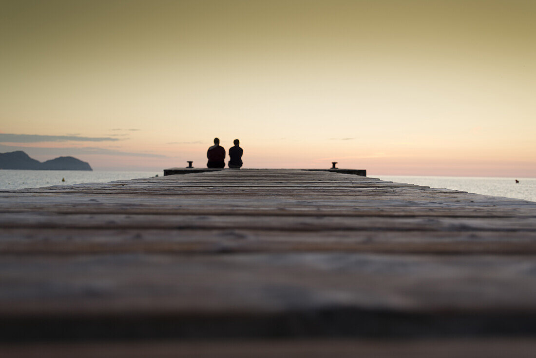 Couple sitting at the end of a long pier in the morning mood. Playa de Muro beach, Alcudia, Mallorca, Balearic Islands, Spain