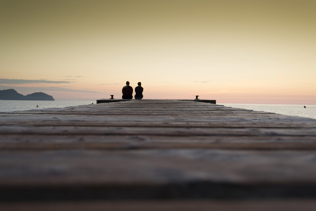 Pärchen sitzt am Ende eines langen Steg in der Morgenstimmung. Strand Playa de Muro, Alcudia, Mallorca, Balearen, Spanien