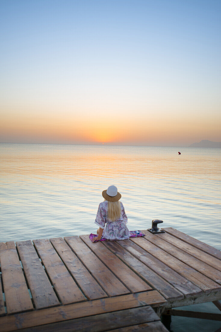 Junge blondhaarige Frau sitzt am Ende eines langen Steg in der Morgenstimmung und genießt den Ausblick auf den Sonnenaufgang. Strand Playa de Muro, Alcudia, Mallorca, Balearen, Spanien