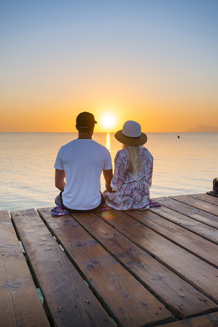 Young couple sitting at the end of a jetty in the morning atmosphere and enjoying the view of the sunrise. Playa de Muro beach, Alcudia, Mallorca, Balearic Islands, Spain