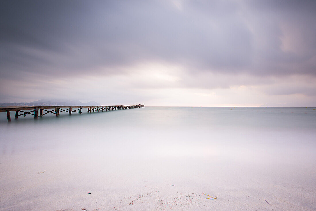 Langzeitbelichtung eines Holzsteg am Strand Playa de Muro in der Morgenstimmung. Der Himmel ist wolkenverhangen und Nebel liegt über der Bucht von Alcudia. Alcudia, Mallorca, Balearen, Spanien