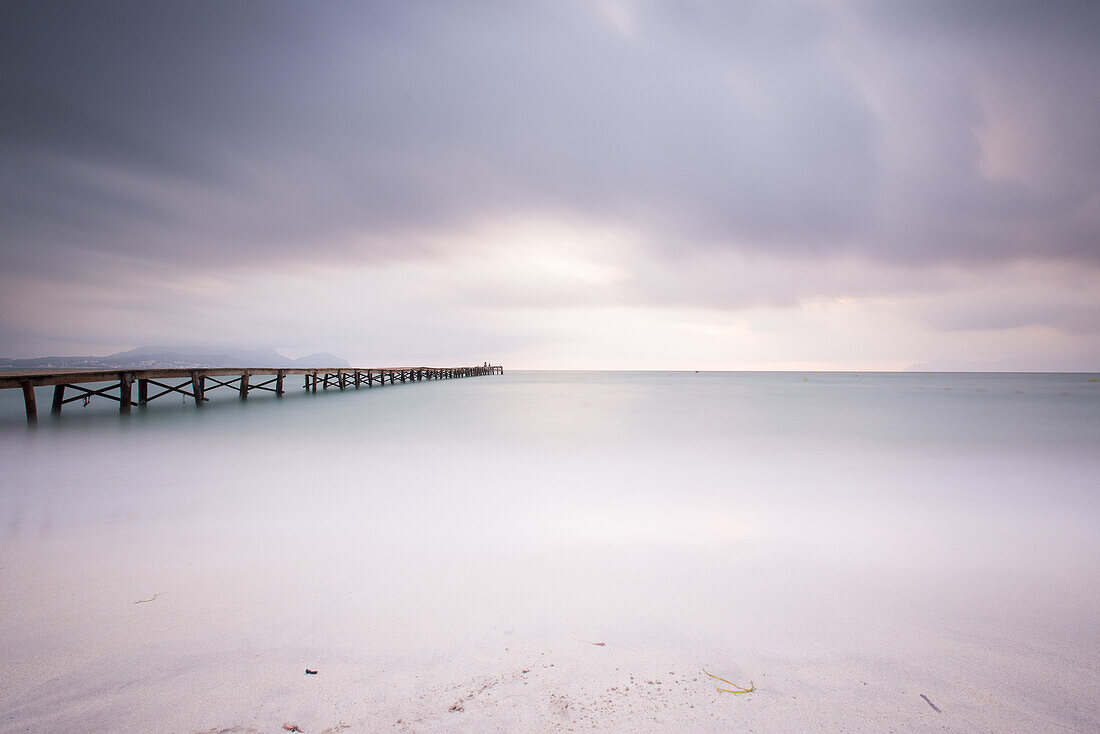 Long exposure of a wooden pier on Playa de Muro beach in the morning mood. The sky is overcast and fog lies over the Bay of Alcudia. Alcudia, Mallorca, Balearic Islands, Spain