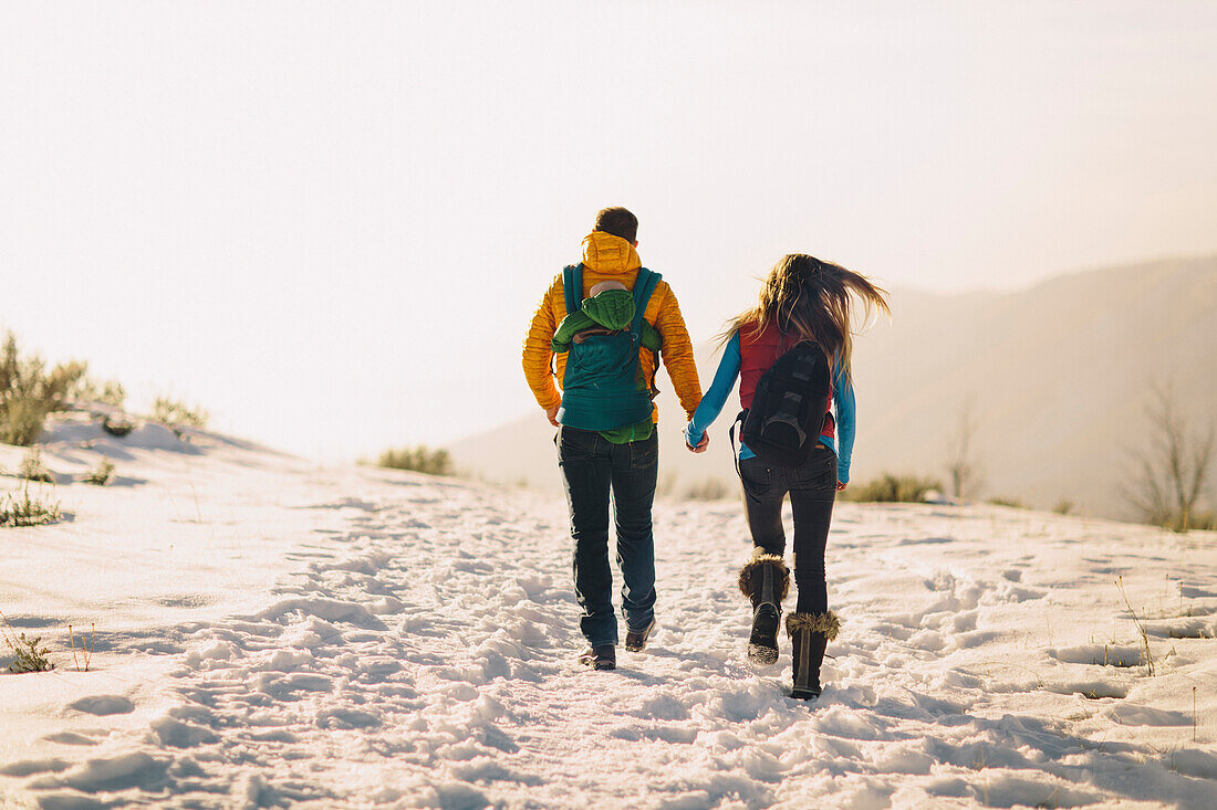 A young couple holds hands and hikes against a winter mountain scene and bright sunset.