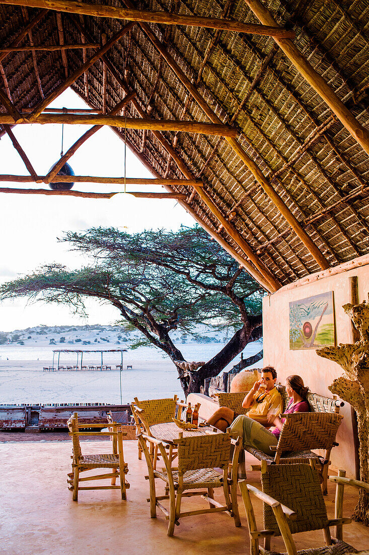 A man and a woman sit in a thatch roof lounge area overlooking an African beach.
