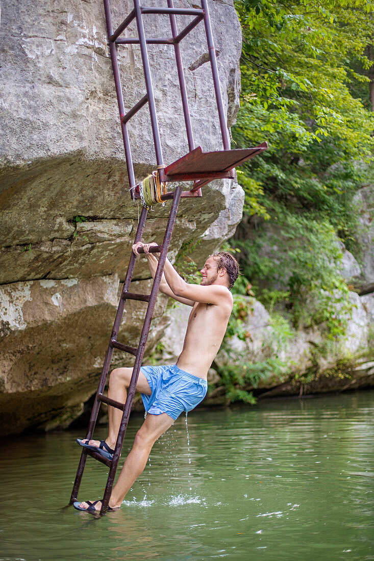 A young man smiles while climbing a ladder on the bank of a river before jumping in.