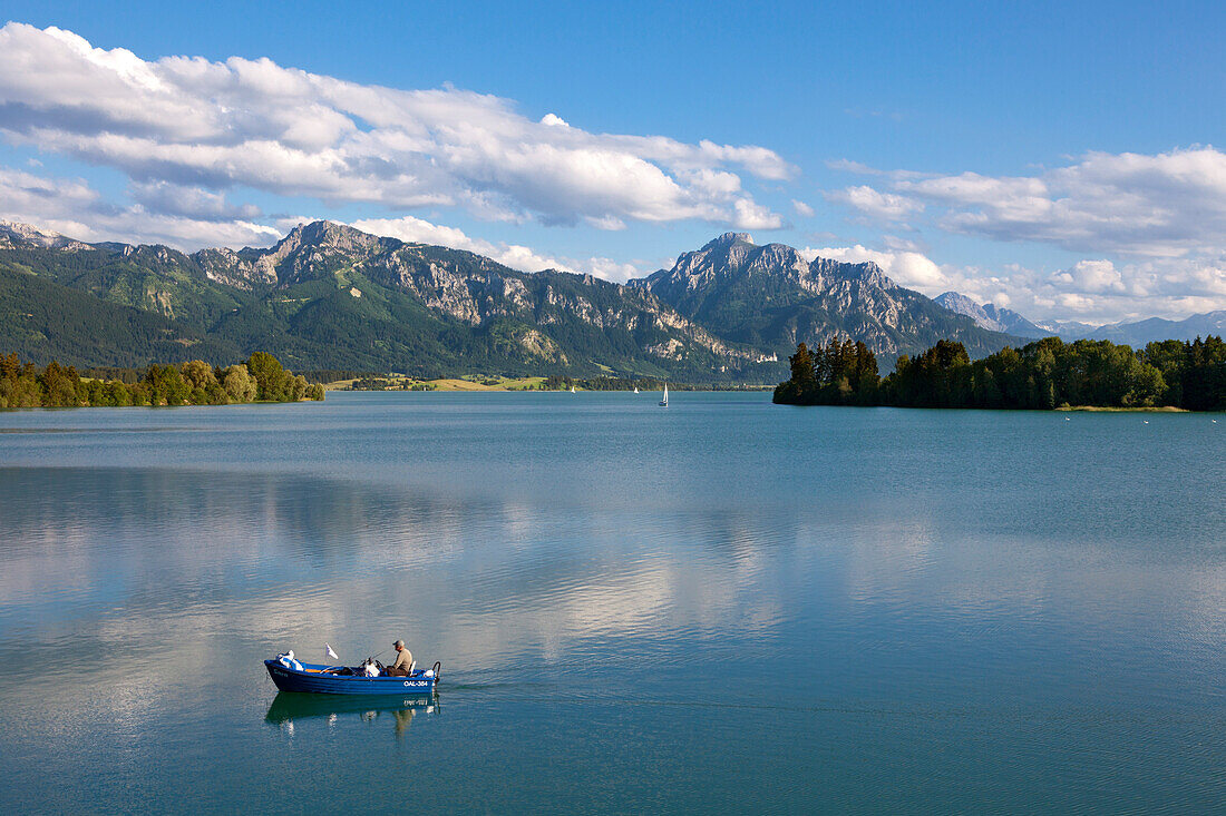 Fisherman in his boat on lake Forggensee, view to Tegelberg, Saeuling and Neuschwanstein castle, Allgaeu, Bavaria, Germany