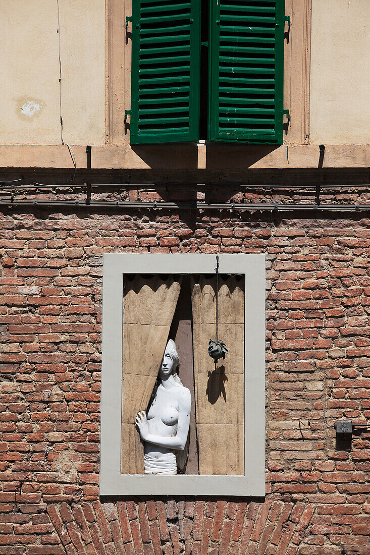 A statue in the window of a brick building, looking out past the curtains, Montepulciano, Tuscany, Italy