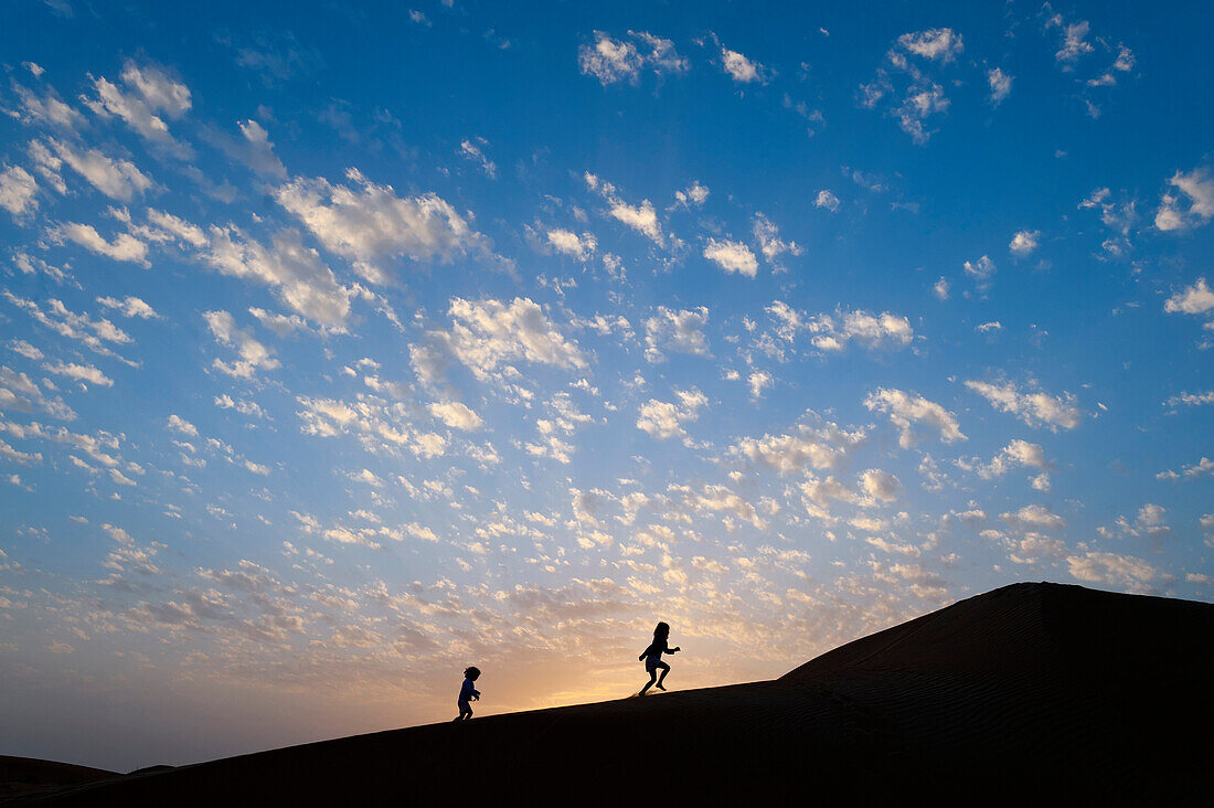 Two young girls running up sand dune at dusk, Dubai, United Arab Emirates