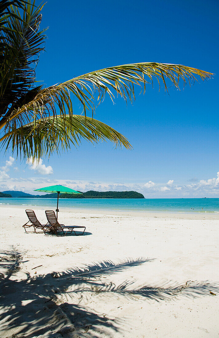 Deck chairs under parasol on white sandy beach with palm trees overlooking blue sea, Cenang beach, Pulau Langkawi, Malaysia