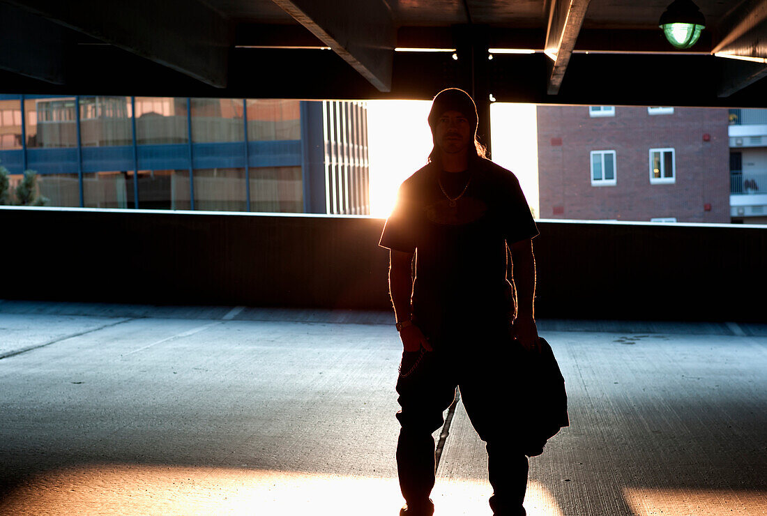 'A young man stands in an elevated parking lot backlit by the setting sun in an urban area; Burlington, Vermont, United States of America'