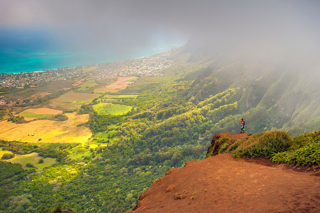 'A man standing on the edge of a cliff on the Kuliouou Ridge Trial enjoys the view of Oahu's windward side and the town of Waimanalo as the clouds roll in on a summer's day; Waimanalo, Oahu, Hawaii, United States of America'