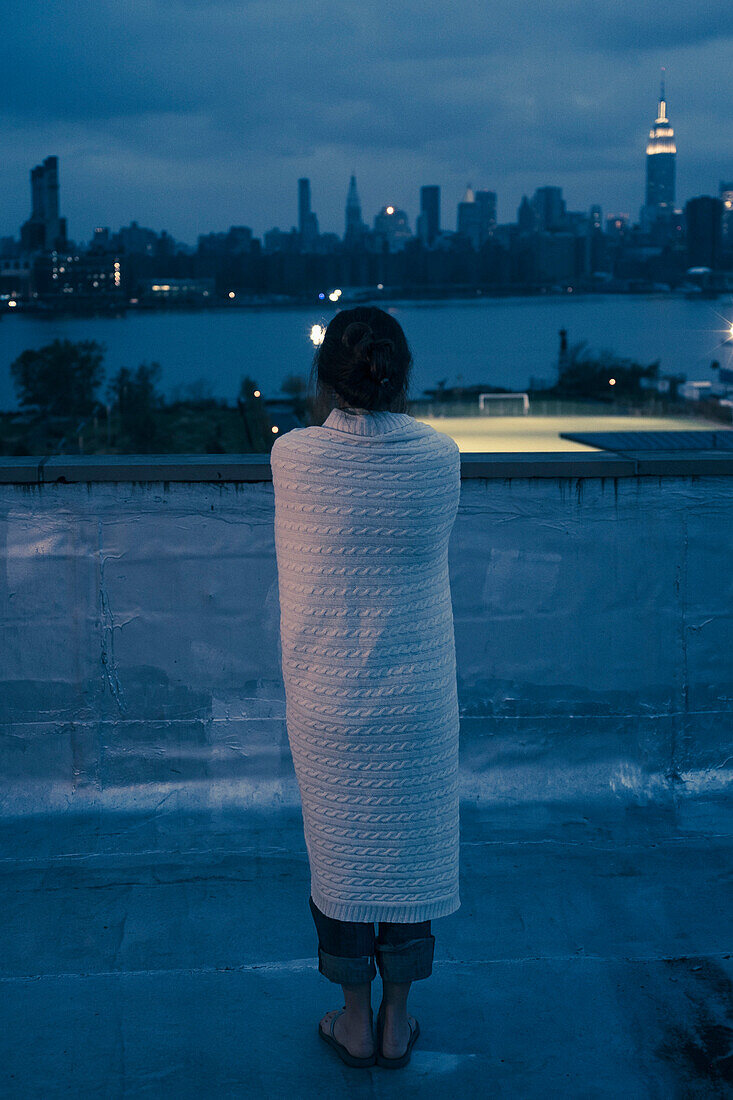 Caucasian woman wrapped in blanket admiring urban waterfront from rooftop