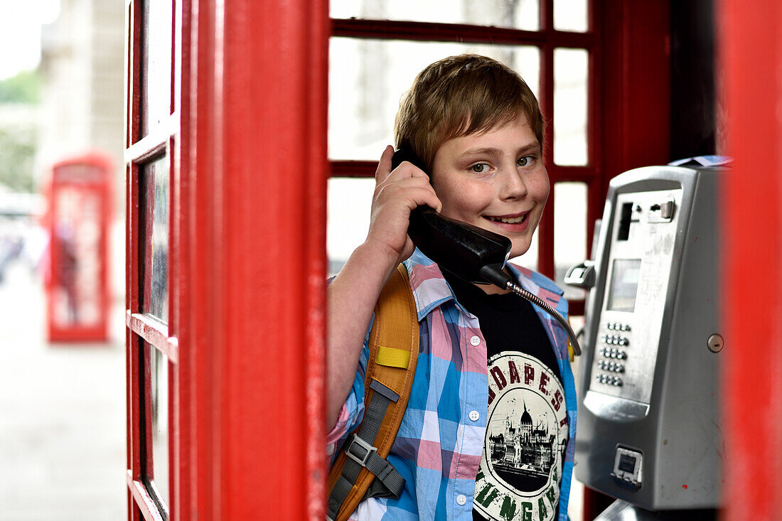 Boy (11) inside a typical British, red phone box with telephone in his hand, London, Great Britain