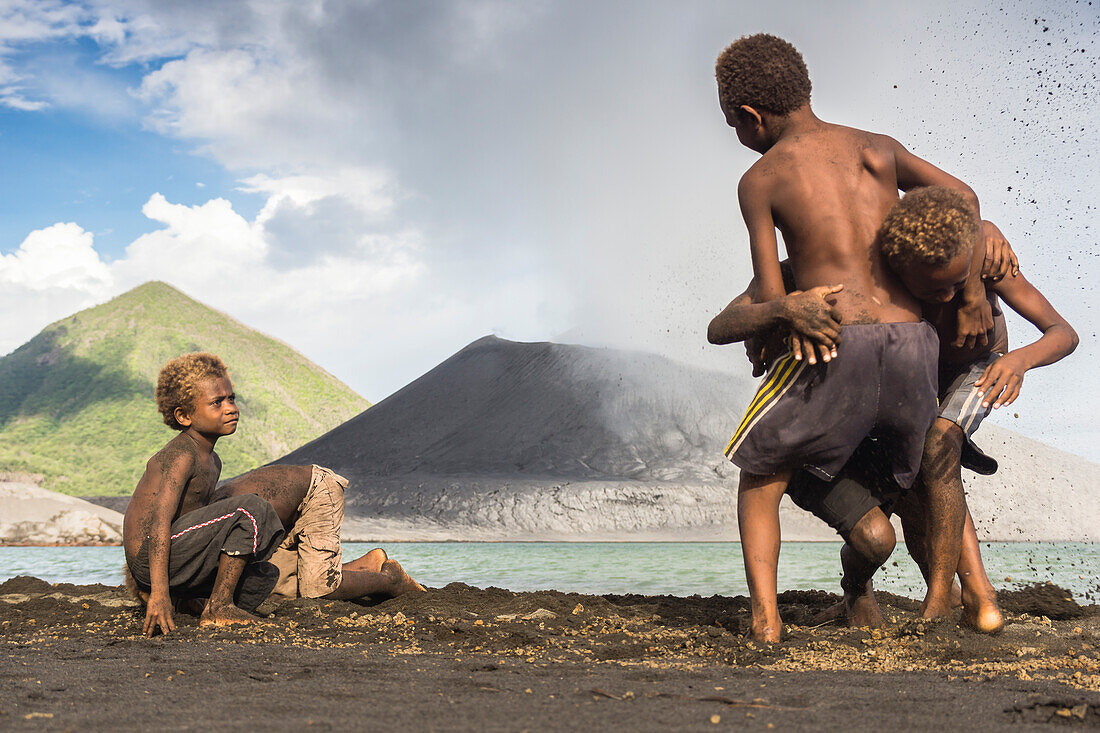 Kinder spielen im Aschesand am Fuße des am Meer gelegenen aktiven Vulkan Tavurvur während einer Eruption. Im Hintergrund der grüne Kegel des zweiten Vulkans ''Vulcan'' zu sehen, Papua Neuguinea