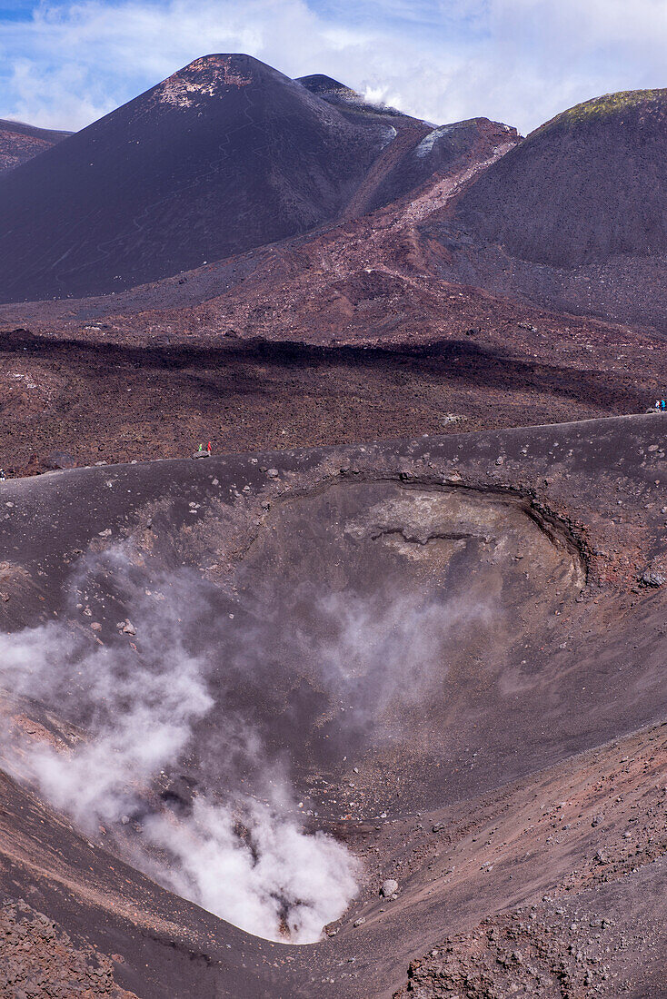 Großer aktiver Vulkankegel im Schatten des Etna mit erstarrtem Lavafluss des Südost-Kraters. Heißer Wasserdampf tritt aus dem Krater. Menschen laufen um den Kraterrand, Vulkan Etna, Sizilien, Italien