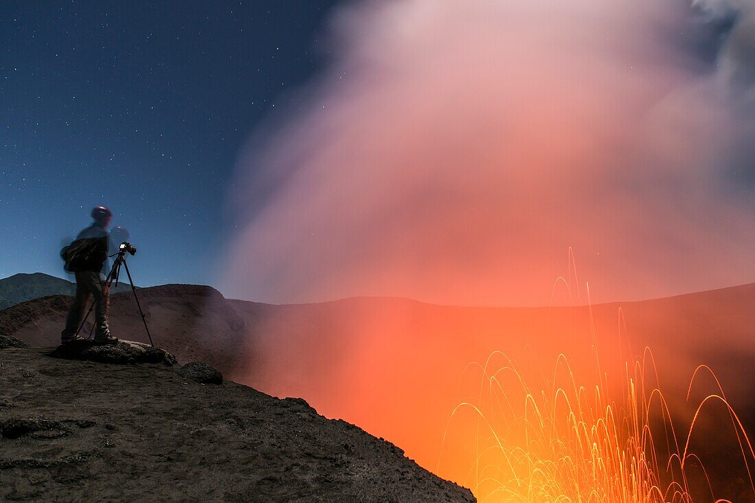 During an eruption, man stands with tripod, camera and helmet on the edge of the volcano Yasur at night with starry sky. Vanuatu, Tanna Island, South Pacific