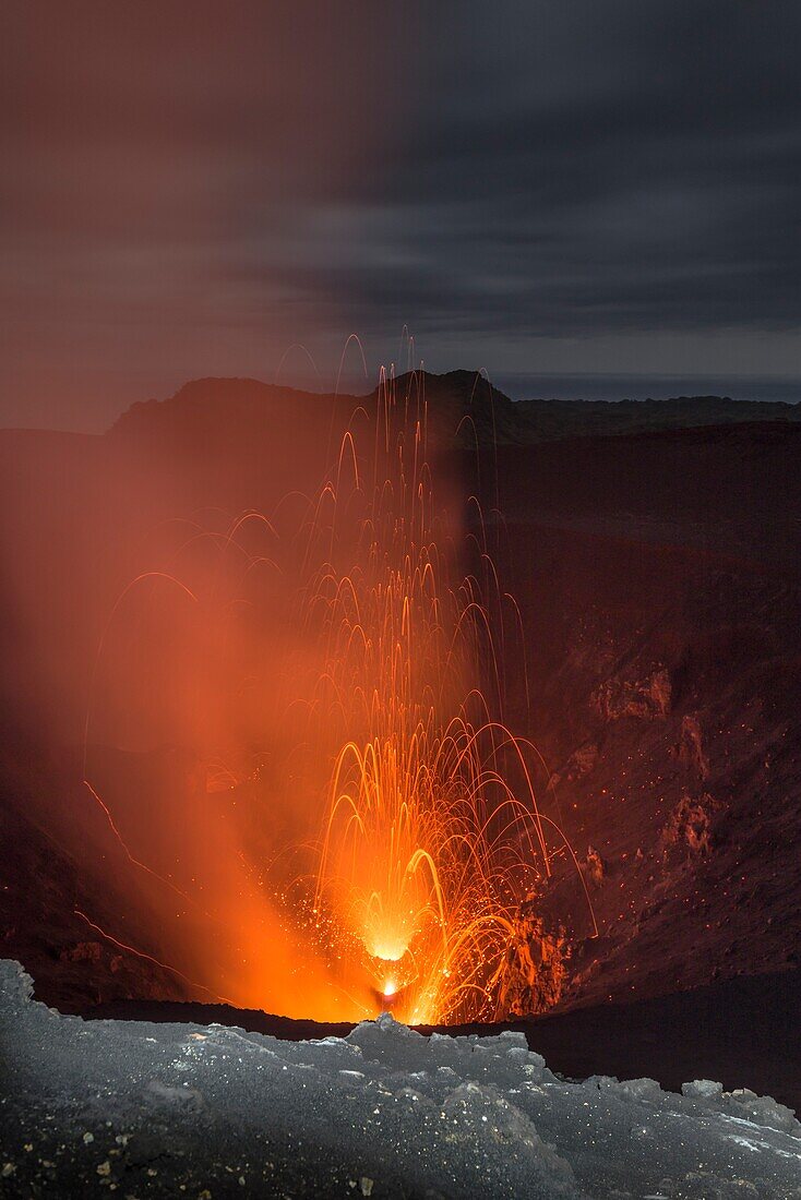 Hohe Lava-Explosion des aktiven Vulkan Yasur mit Lava-Bomben und giftigen Gasen. Der Kraterrand im Vordergund ist mit einer Taschenlampe aufgehellt, bedeckter, dunkler Himmel zur der blauen Stunde, Vanuatu, Insel Tanna, Süd Pazifik