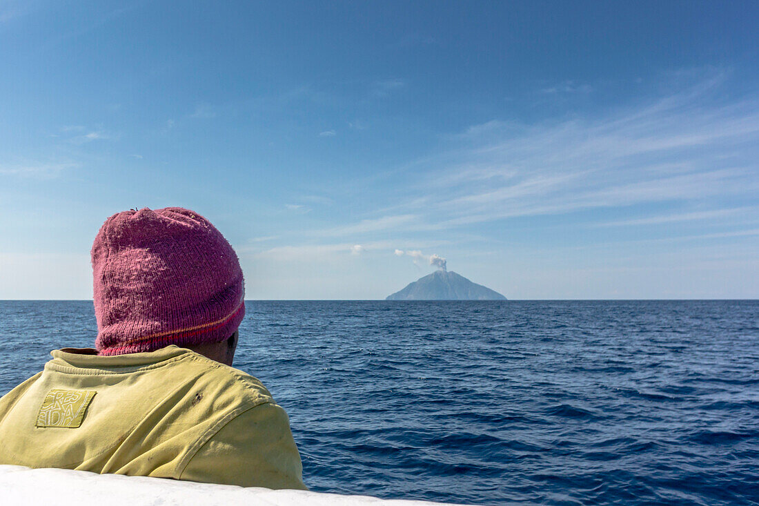 Man sitting in a boat in the open sea,  looking towards the cone of Batu Tara volcano (burning stone), , island of Komba, Flores Sea, Indonesia