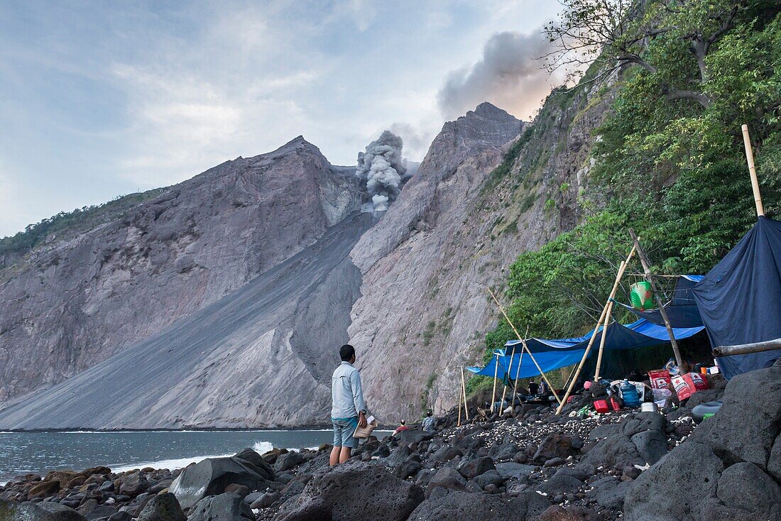 Base Camp at the foot of the active volcano Batu Tara between the sea and cliffs. Man shocked by an eruption, Island Komba, Flores Sea, Indonesia