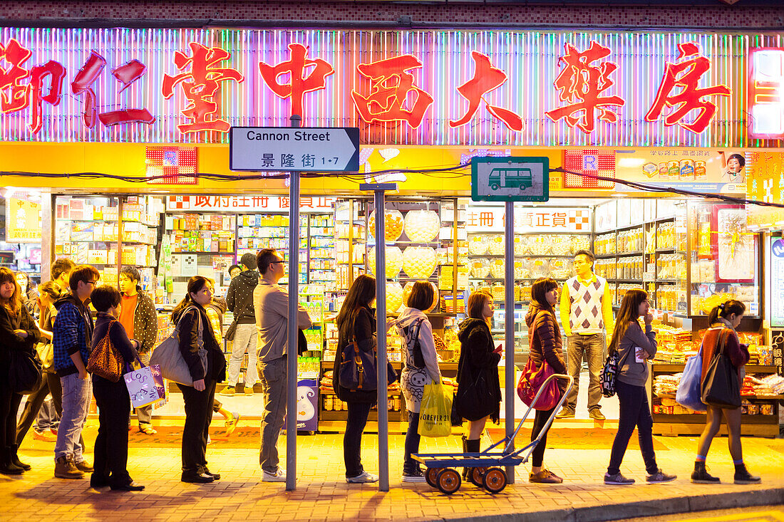Passanten stehen an einer Haltestelle an, Straßenbahn, vor einer Apotheke, abends, Leuchtreklame, Bürgersteig, Einkaufsgegend Causeway Bay, Schilder, Werbung, chinesische Schriftzeichen, Straßenszene, Hongkong, China, Asien