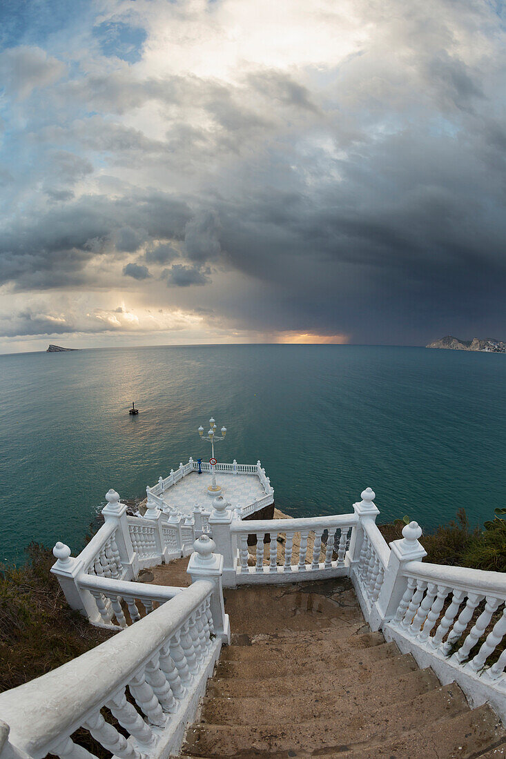 Steps leading down to the turquoise water with the sun glowing on the horizon under storm clouds, Benidorm, Spain